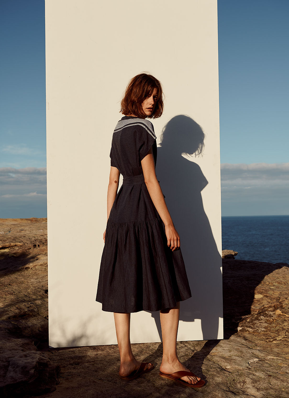 A knee-length dress with cap sleeves, gathered detailing and tie belt of the same fabric made from yarn dye stripe washed linen in ink with pale blue stripe