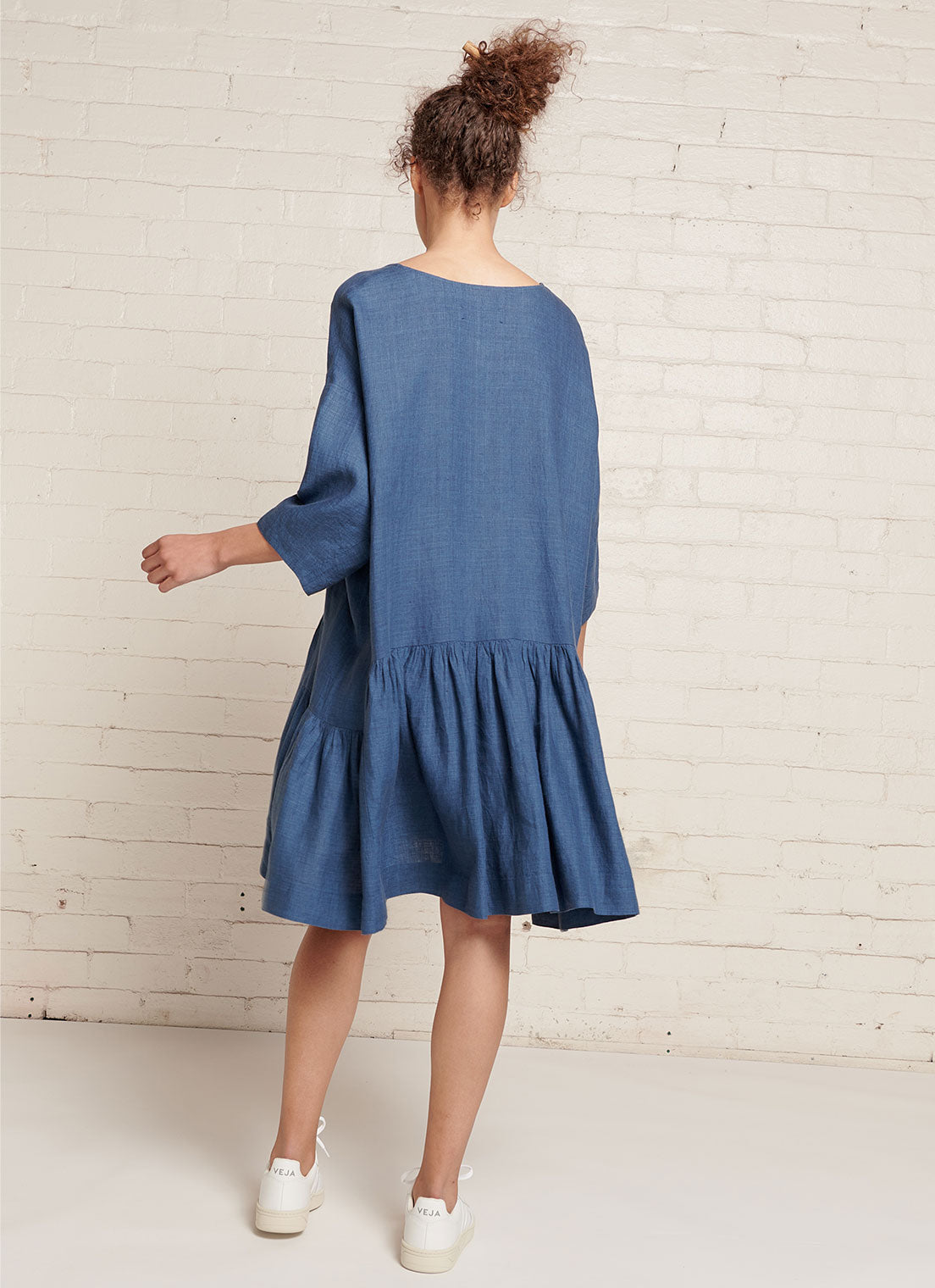 An indigo, easy fit, knee-length, square-cut dress with open neckline, 3/4 sleeves, and gathered detailing made from yarn dye washed linen