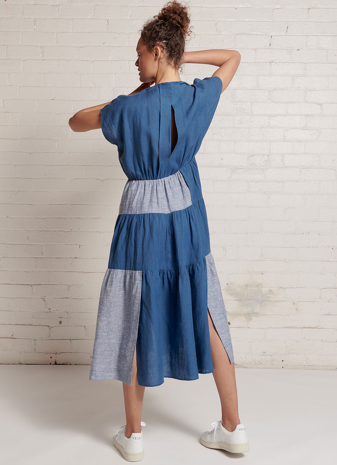 An indigo and denim two-tone, patchwork, midi tiered dress with capped sleeves, pleated detail on the neckline and at the back, elastic waist and slits on the skirt made from yarn dye washed linen