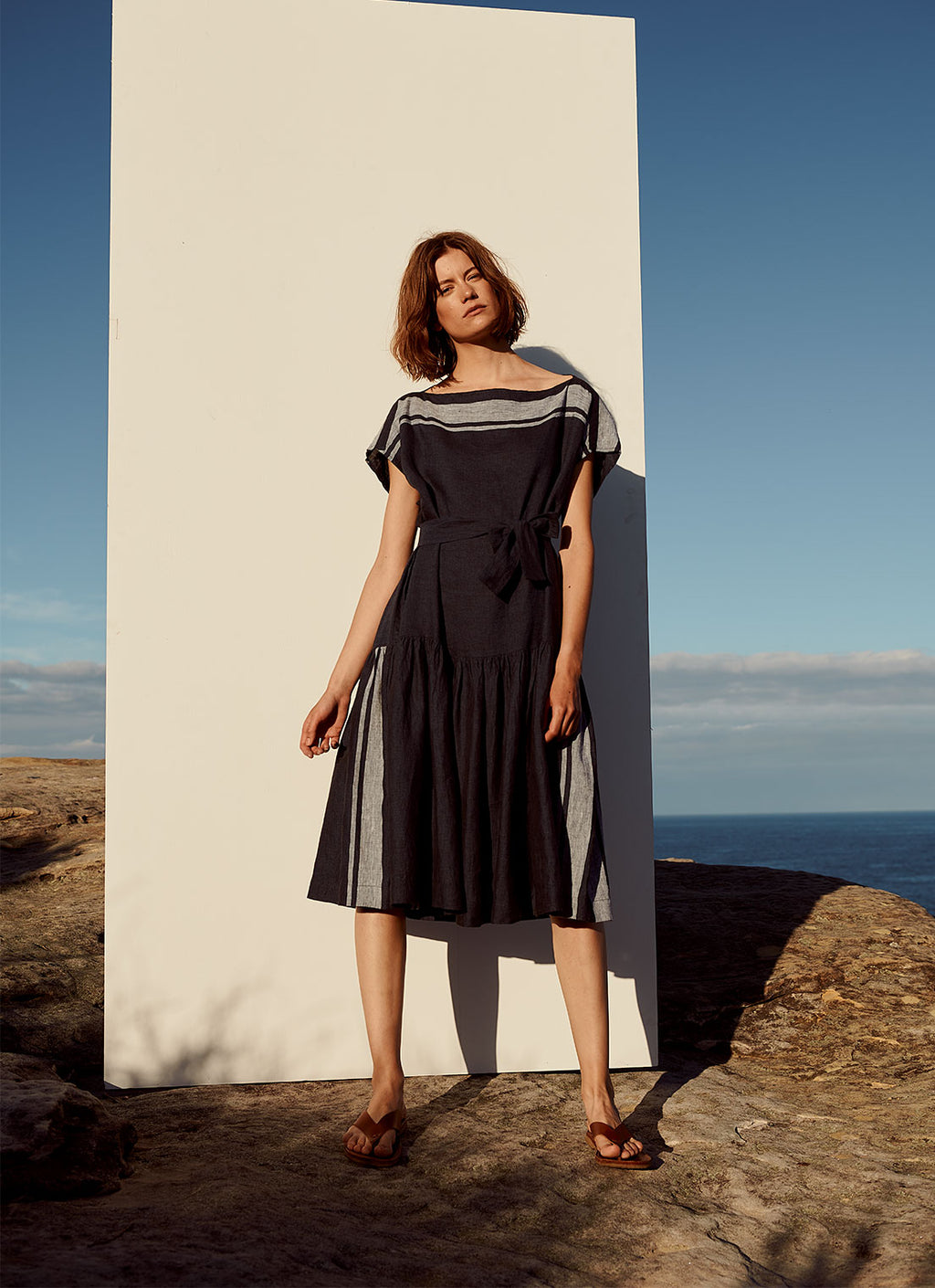A knee-length dress with cap sleeves, gathered detailing and tie belt of the same fabric made from yarn dye stripe washed linen in ink with pale blue stripe