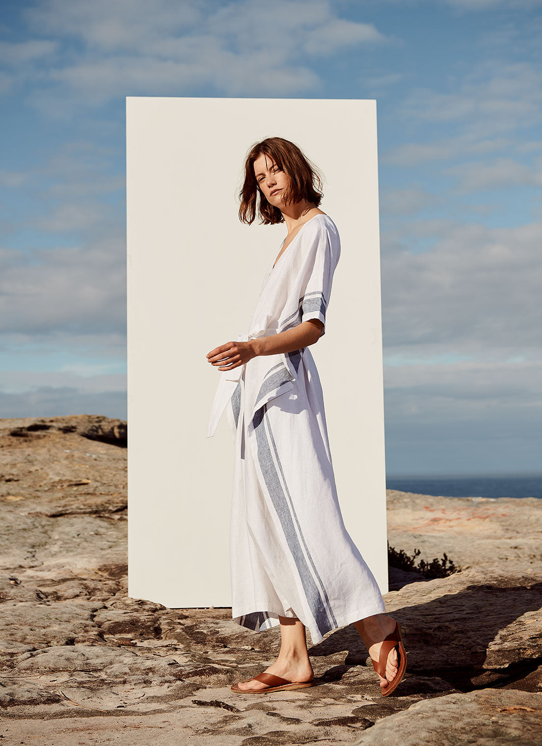 A rectangular cut, v-neck jumpsuit with short sleeves, centre front button closure and tie belt of the same fabric made from yarn dye stripe washed linen in white with indigo stripe
